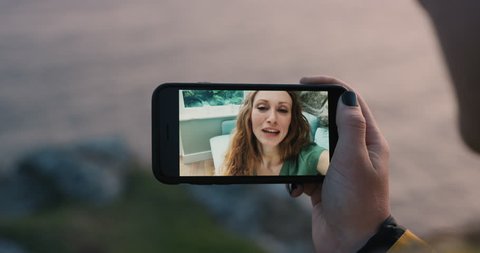 Young woman having video chat holding smartphone webcam chatting to attractive redhead girlfriend outdoors by the ocean at sunset