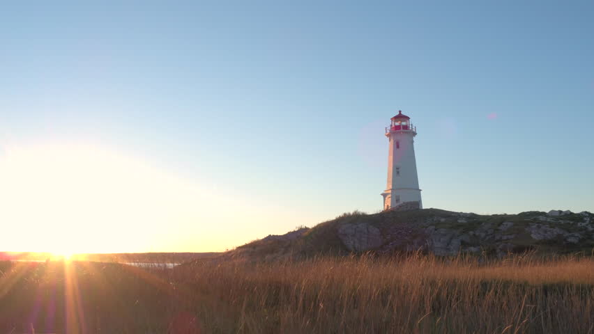 Beautiful Louisbourg Lighthouse standing on rocky and grassy coast rising above North Atlantic Ocean on Nova Scotia peninsula, Canada at golden light sunset. Sunrays illuminating dry straw on seashore