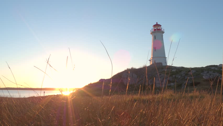 Beautiful Louisbourg Lighthouse standing on rocky and grassy coast rising above North Atlantic Ocean on Nova Scotia peninsula, Canada at golden light sunset. Sunrays illuminating dry straw on seashore