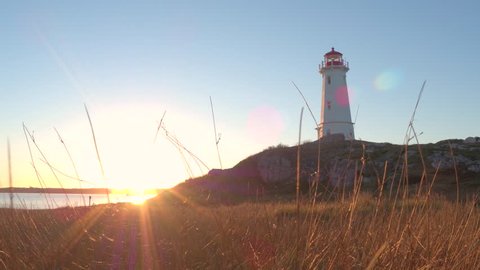 Beautiful Louisbourg Lighthouse standing on rocky and grassy coast rising above North Atlantic Ocean on Nova Scotia peninsula, Canada at golden light sunset. Sunrays illuminating dry straw on seashore