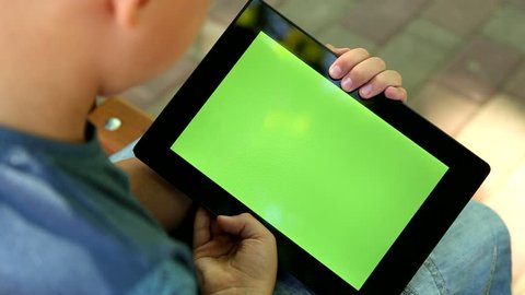 Closeup of handsome blond boy sits on bench in summer or autumn city park. Boy looking at blank green screen of modern tablet pc. Real time full hd video footage. View from above.