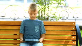 Cute smiling blond boy sits on bench in summer or autumn city park with blurry moving automobiles in background. Boy looks at blank green screen of modern tablet pc. Real time full hd video footage.