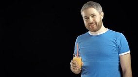 Athletic bearded young man in blue tshirt drinking orange juice with a straw. Black background. 4K video