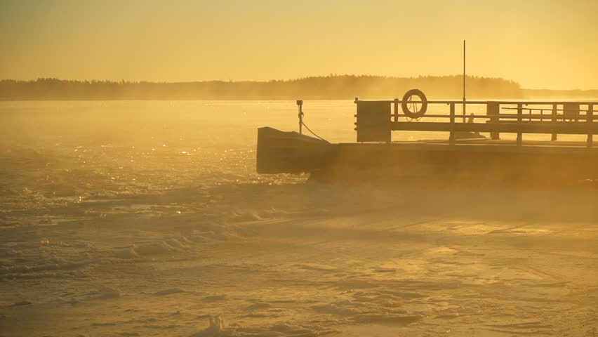 snowstorm and blizzard on frozen shining lake with thawing ice and waving water with melting floes near the pier under the sunset light during winter cold weather