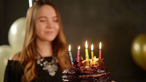 Cute Young Girl with Long Hair is Blowing Candles on a Chocolate Cake Standing on Black Background with Golden and White Air Balloons. Birtday Girl and Sweet Dessert.