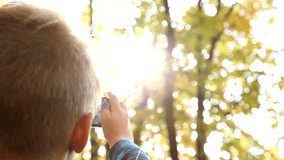 Close up of back of happy little child and smartphone in hands. Blond caucasian boy takes picture of beautiful landscape with bright foliage and sunlight using modern mobile phone with touchscreen.