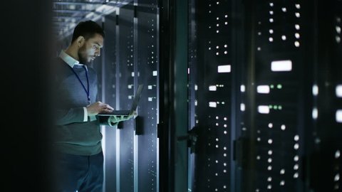Male IT Technician Working on a Laptop Standing Before Open Server Rack Cabinet in Big Data Center. Shot on RED EPIC-W 8K Helium Cinema Camera.