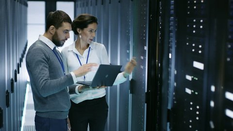 Male IT Specialist Holds Laptop and Discusses Work with Female Server Technician. They're Standing in Data Center, Rack Server Cabinet is Open. Shot on RED EPIC-W 8K Helium Cinema Camera.