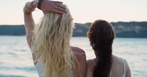Two woman arm in arm watching sunset over lake best friends looking to the future hair blowing in wind wearing denim summer shorts on road trip adventure