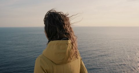 Close up portrait of Young Woman with Red hair blowing in wind looking at sunset over ocean Girl wearing yellow raincoat trekking in Scotland Slow Motion