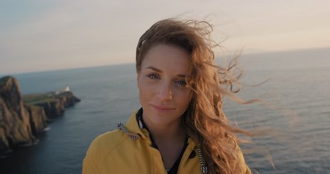 Independent Woman smiling with Red hair blowing in wind Close up portrait looking at sunset over ocean Young Girl wearing yellow raincoat trekking in Scotland Slow Motion