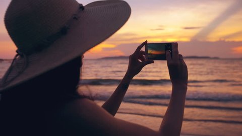 Silhouette of young tourist woman in hat taking photo with cellphone during sunset in ocean beach