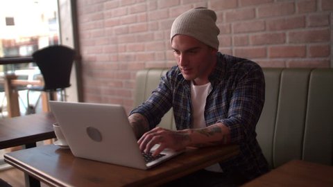 Cool man in casual wear and beanie working on his laptop at modern coffee shop in city