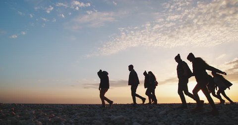 Group of hipster people walking together on a beach with pebbles in winter at sunset