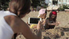 Woman shooting a video of two little cute girls playing together with sand on the beach