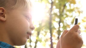 Close up of profile of cute blond boy of 9 years old sitting on bench in city park. Caucasian young boy uses smartphone outdoors over blurry trees background and sunlight. Real time full hd video.