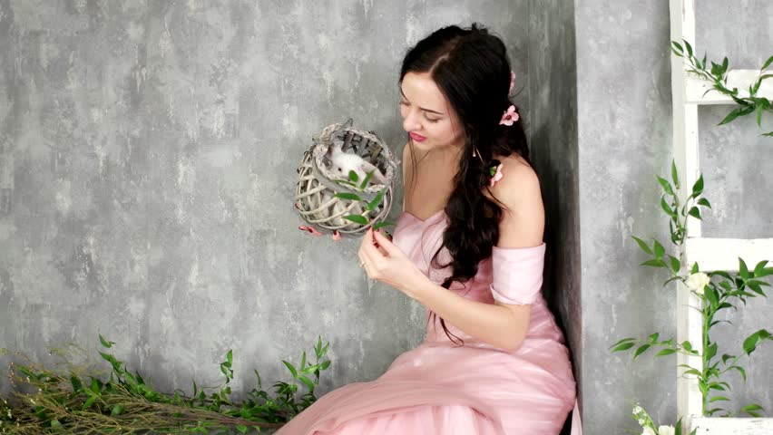 young lady posing in photo studio holding small white rabbit in wicker grey basket, girl having fun with domestic rabbit in photo studio, green plant and flower decoration of grey photo studio