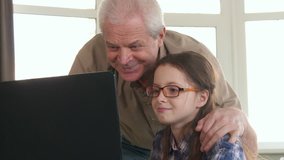 Little qirl in glasses and her grandpa having video chat on laptop. Senior gray man and his gradndaughter waving their hands to the computer screen. Aged caucasian male holding his hand on his