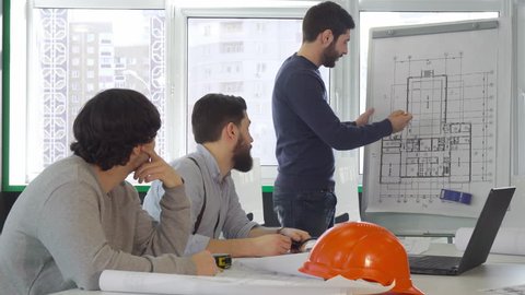 Young architect team discussing project of the building at the office. Brunette bearded man standing near the flip chart. Two caucasian men advicing something to their colleague
