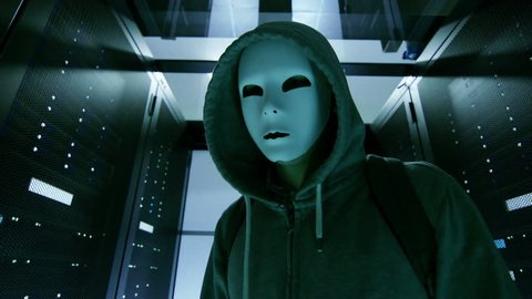 Masked Hacker in a Hoodie Walks Through Corporate Data Center with Rows of Working Rack Servers. Shot on RED EPIC-W 8K Helium Cinema Camera.