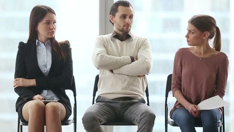 Group of three young applicants sitting on chairs in modern office lobby, waiting for job interview, feeling nervous. Funny rivals looking at each other with arrogant face expressions. Job search