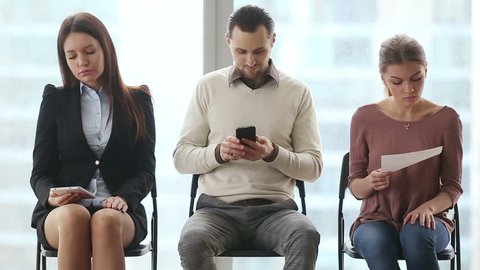 Group of three young casual caucasian candidates sitting on chairs in modern office lobby, waiting for job interview, feeling nervous. Guy talking on phone about his competitors. Job search concept
