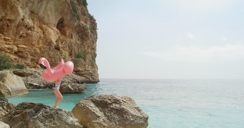 Woman walking over rocks on beach carrying inflatable flamingo enjoying summer vacation on tropical island holiday