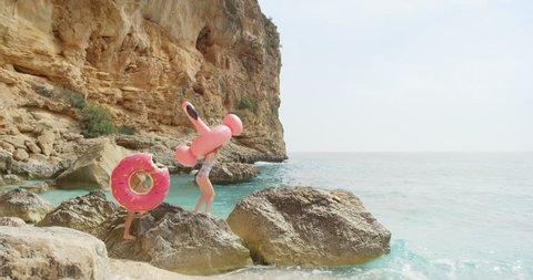 Two girls walking over rocks on beach carrying inflatable flamingo and doughnut Best friends women enjoying summer vacation on tropical island holiday