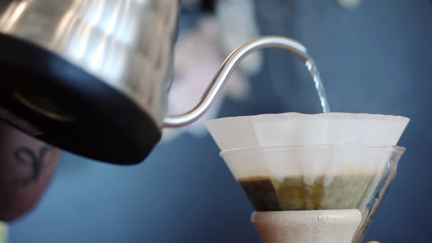 He pours hot coffee water from a metal kettle, which is poured into a paper filter. Coffee foams.