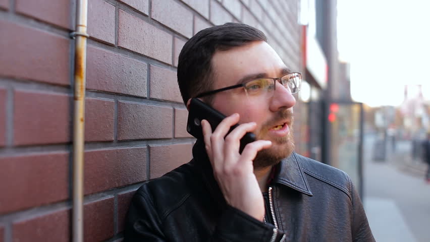 Young lonely man talking on the phone near the brick wall in the city