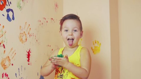 Little boy in yellow sleeveless shirt showing tongue. His hands are dirty in colors. He is living his handprints on the wall. Slowmotion