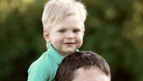 Cute baby of 1 year old sits on shoulders of father. Little caucasian blond child and daddy plays active games outdoors in summer park. Real time full hd video footage.