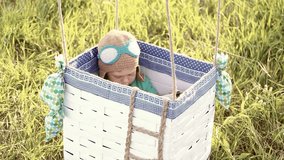 Beautiful funny little child wearing knitted pilot hat sits in handmade toy airballoon in green grass. Portrait of cute baby looking at camera smiling happily. Real time full hd video clip.