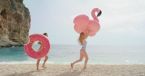 Two women walking on empty beach holding giant inflatable flamingo and doughnut Best friend girls enjoying summer vacation on tropical beach holiday