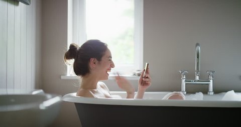 Beauty woman lying in bubble bath having video chat using smartphone webcam chatting to friend at home