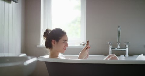 Beautiful Woman relaxing in bubble bath lying in bathtub using smartphone browsing social media sharing photos mindfulness health concept