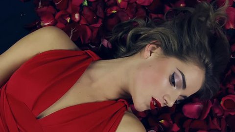 Girl in red dress with beautiful hairdo lies in petals red roses,slowly turns head from the side, hands touch her face.Valentine's Day.Girl in love.Red lipstick and bright makeup.Background of flowers