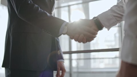 Low angle shot with tilt up of two businessmen in official clothes greeting each other with handshake and discussing work before big window