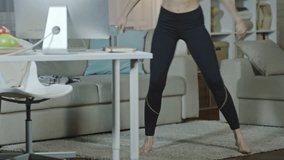 Tilt up of young sporty woman watching online fitness course and doing side lunges in the living room and then walking to computer and typing on keyboard