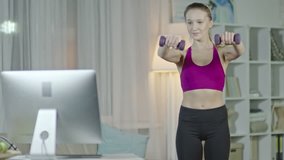 Tracking shot of young woman watching workout online class on computer and doing front and lateral dumbbell raises at home