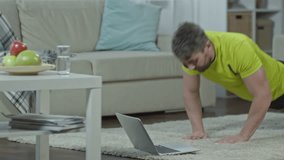 Slow motion shot of strong bearded man doing pushups on the floor in living room and watching workout video tutorial on laptop