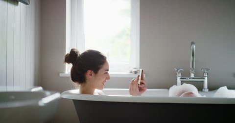 Beautiful Woman relaxing in bubble bath lying in bathtub using smartphone browsing social media sharing photos mindfulness health concept