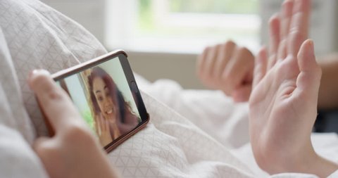 Young woman having video chat holding smartphone webcam chatting to girlfriend lying in bed at home
