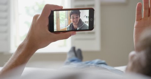 Young homosexual man having video chat holding smartphone webcam chatting to boyfriend lying in bed at home