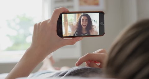 Young woman having video chat holding smartphone webcam chatting to girlfriend lying in bed at home