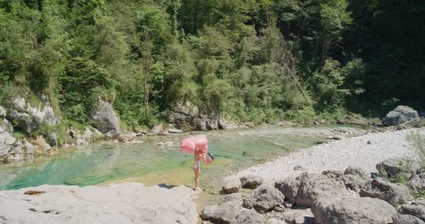 Crazy Young woman carrying inflatable flamingo walking along river Beautiful girl searching for paradise in crystal clear blue water on bright sunny summer day enjoying nature wild outdoors