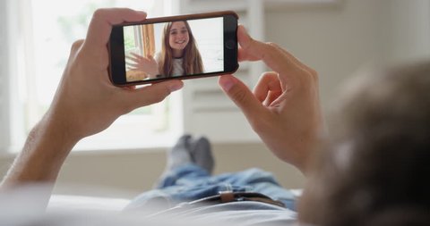 Young Man having video chat holding smartphone  webcam chatting to young teenage girlfriend lying in bed at home