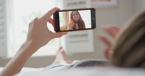 Young woman having video chat holding smartphone  webcam chatting to girlfriend lying in bed at home