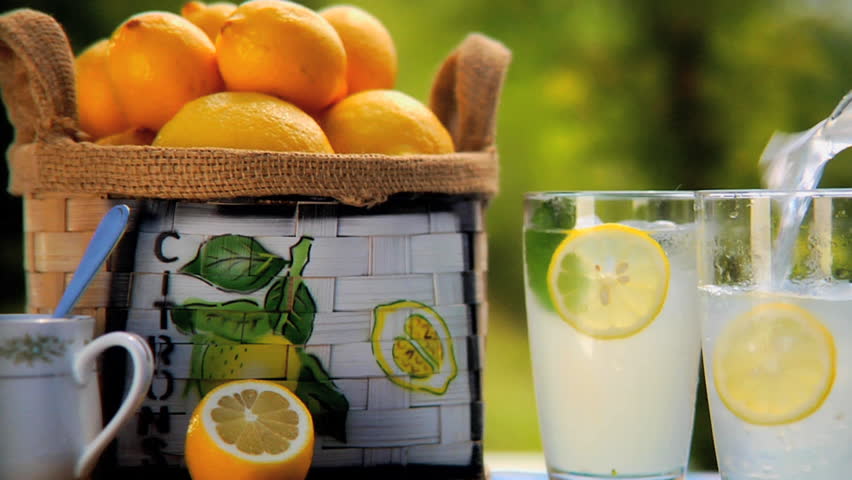 Pan over basket of lemons and man pours 2 glasses of lemonade on table outside