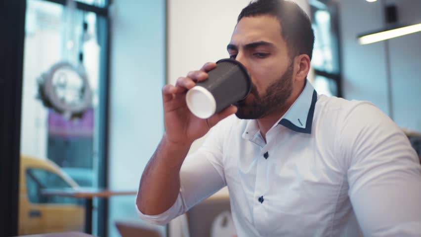 Handsome athletic man having a cup of coffee, while emailing with his phone, modern cafe. Social networks, being online. Modern cafe interior, no people around.